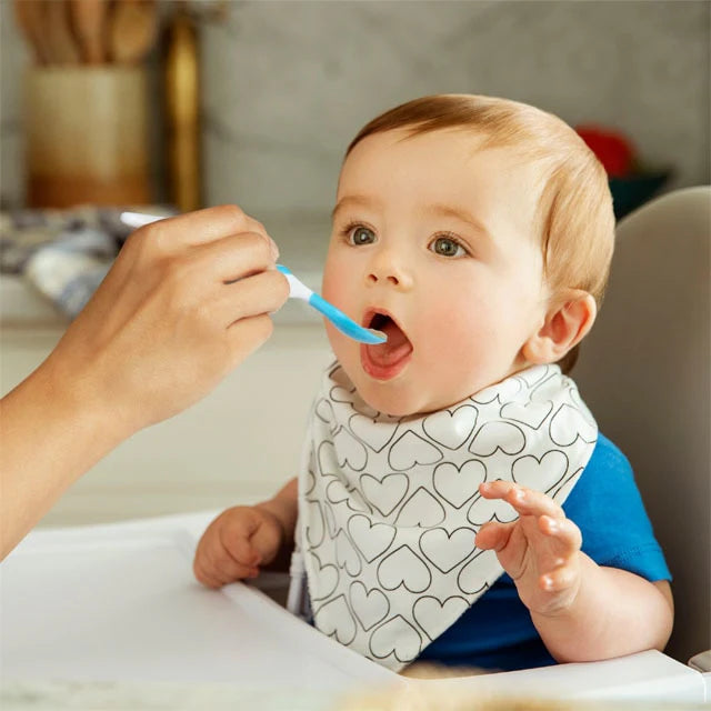 Baby being fed with a spoon by an adult, wearing a bib with heart patterns.